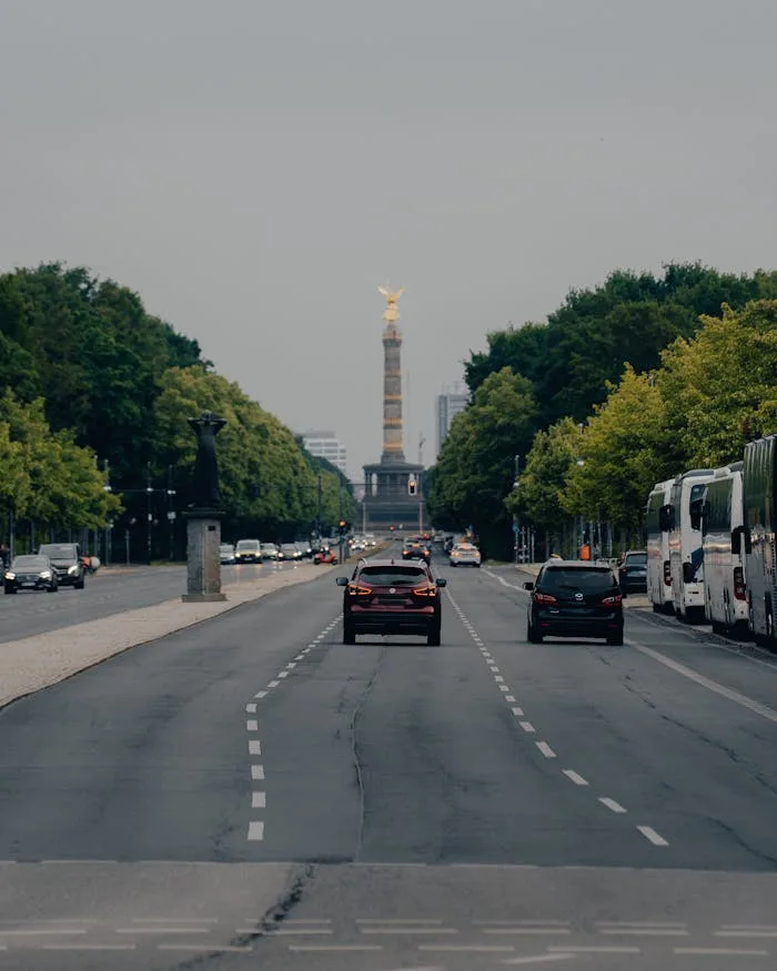 hero-img-02 Cars driving towards the Victory Column on a broad avenue in Berlin, surrounded by lush green trees.